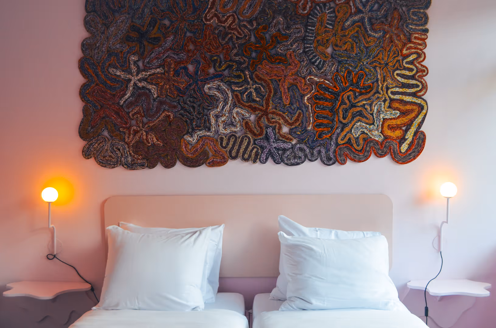 Twee eenpersoonsbedden met witte lakens in een kunstzinnige hotelkamer van De Groene Afslag, met een kleurrijk wandtapijt en zachte verlichting aan beide zijden van het bed.