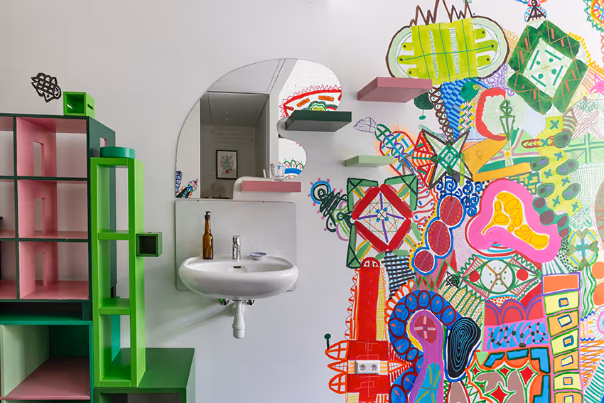Witte kamer met een wit wastafel, een onregelmatige spiegel erboven, groene en roze open kast links en een kleurrijke abstracte muurschildering rechts.