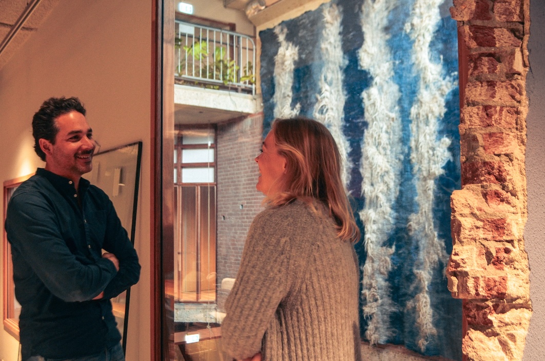 Twee mensen die lachen en met elkaar praten in een interieur met een muur van baksteen en een kunstwerk van blauwe en witte wol.