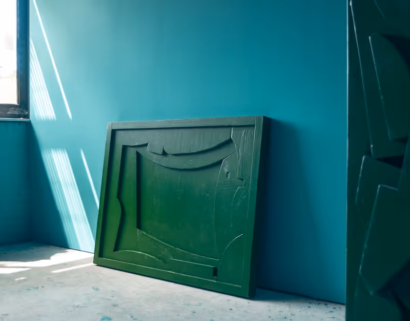 Groene houten kunstwerk met abstract patroon leunt tegen een blauwe muur bij een raam met zonlicht.