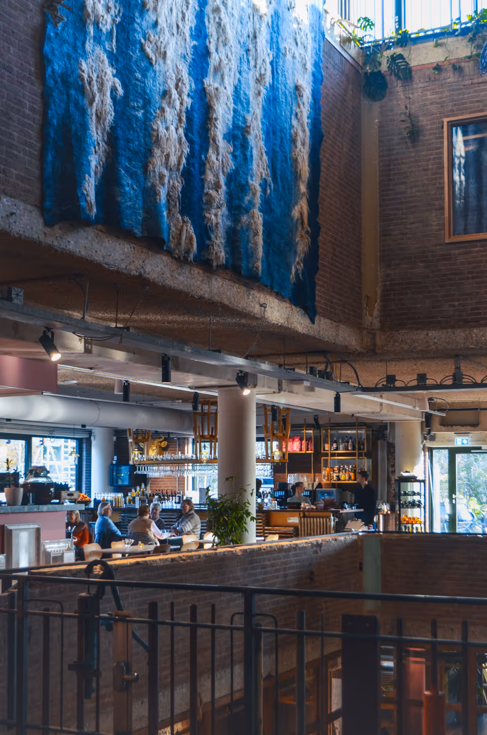 Sfeerbeeld van het restaurant van De Groene Afslag in Bussum. Aan tafeltjes zitten mensen te eten, boven hen hangt een groot blauw kunstwerk van textiel. 