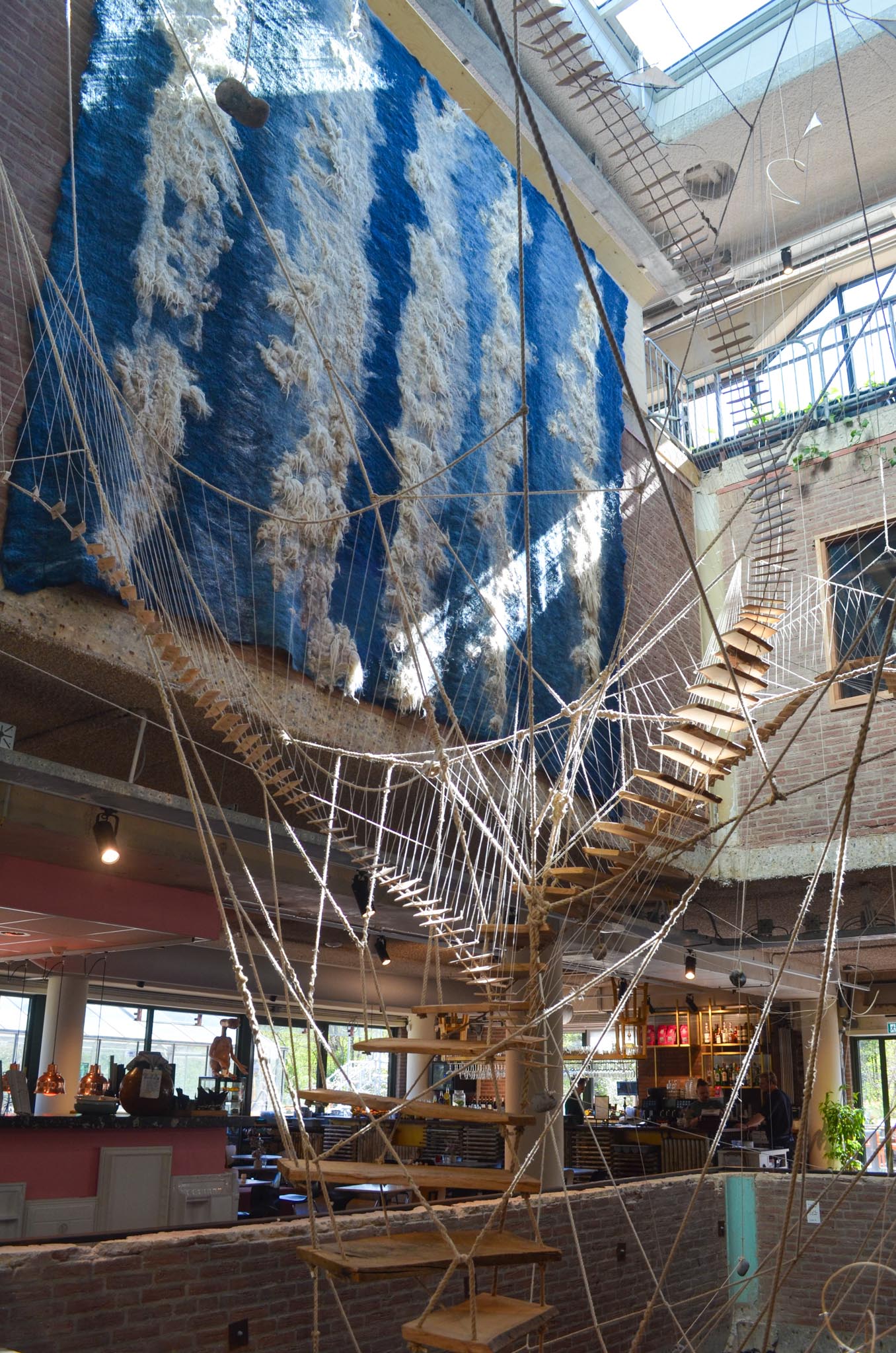 Interieur van een modern gebouw met een kunstinstallatie van houten trappen verbonden door touwen, hangend onder een groot dakraam.