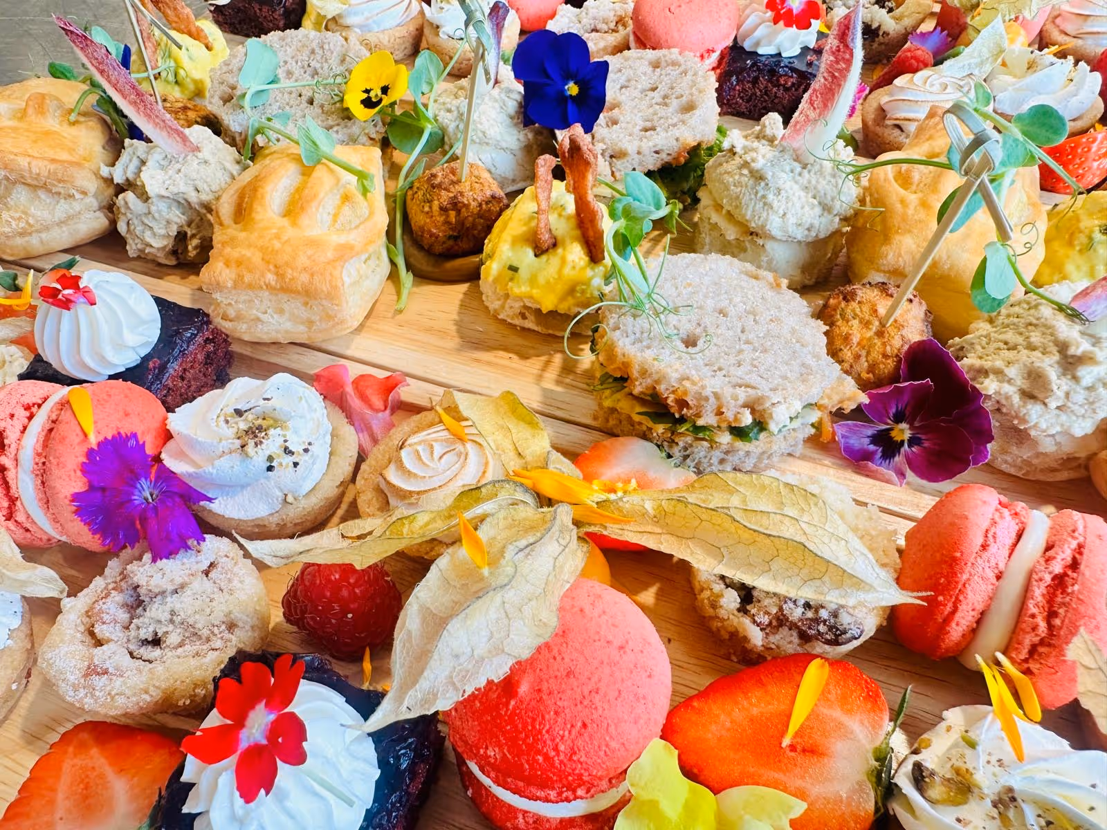 Diverse kleurrijke mini sandwiches en zoete gebakjes versierd met bloemen en fruit op een houten plank.