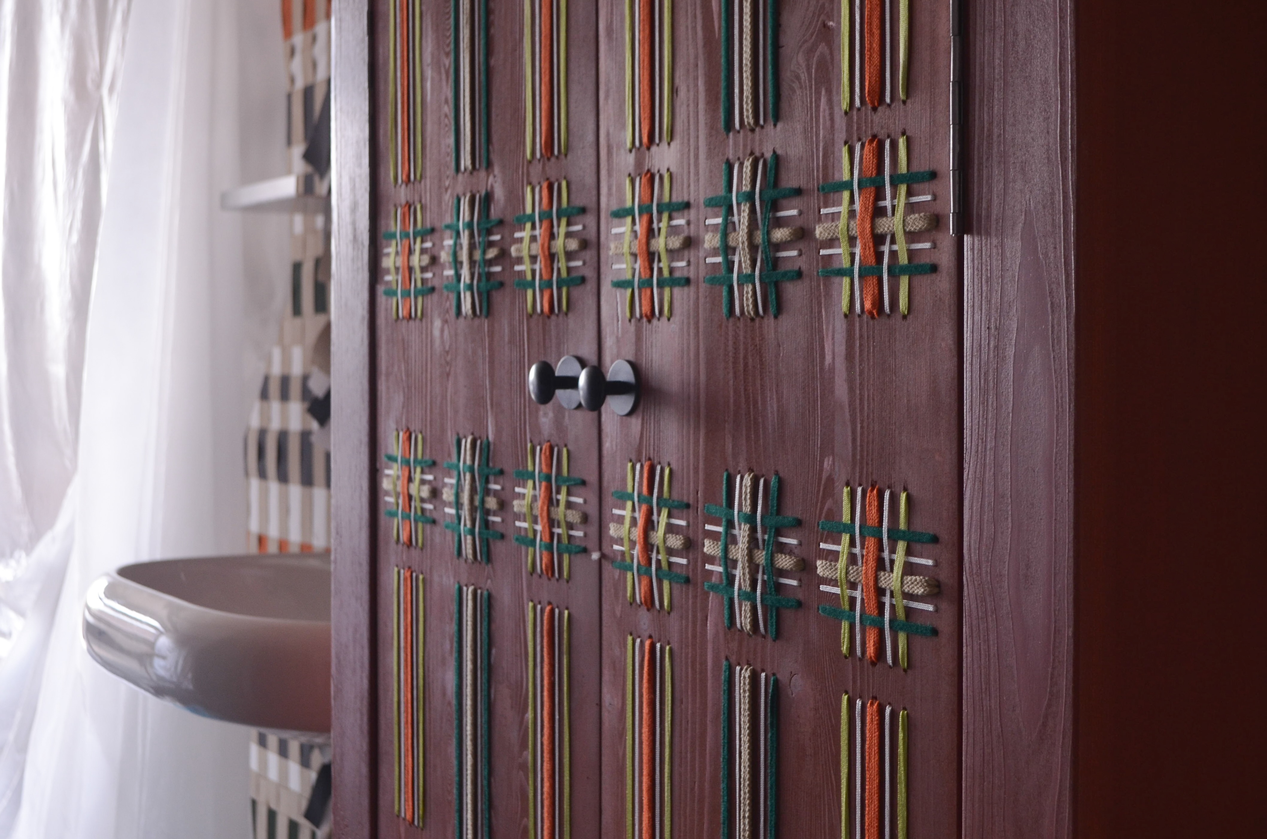 Detail van houten kastdeuren in de Dreamweaver kunstkamer van hotel De Groene Afslag, versierd met kleurrijk geweven ruitpatroon van textiel in groen, oranje en beige. Op de achtergrond een wasbak en een wit gordijn in een sfeervolle hotelkamer.