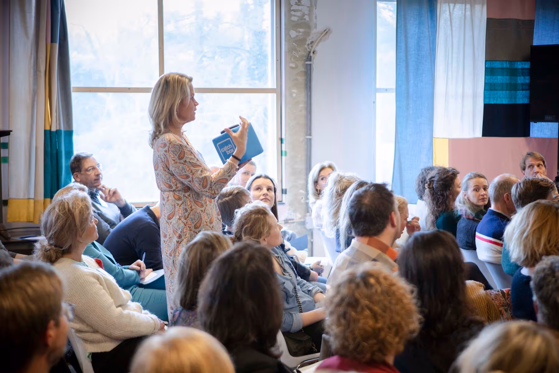Vrouwelijke spreker met een blauwe Catchbox-microfoon in haar hand staat in een zaal tussen zittend publiek dat aandachtig naar haar luistert.