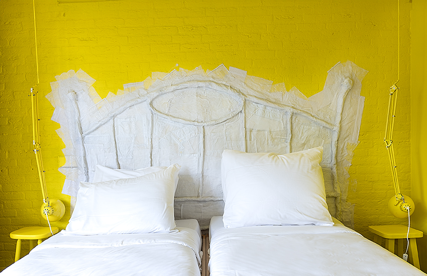 Wanddetail in de nog in aanbouw zijnde hotelkamer “This Room Has Moved” van hotel De Groene Afslag, met een felgele muur en een onaf kunstwerk dat lijkt op een bedhoofdbord. Op de felblauwe vloer staan twee gele krukjes nog ingepakt in bubbelplastic.