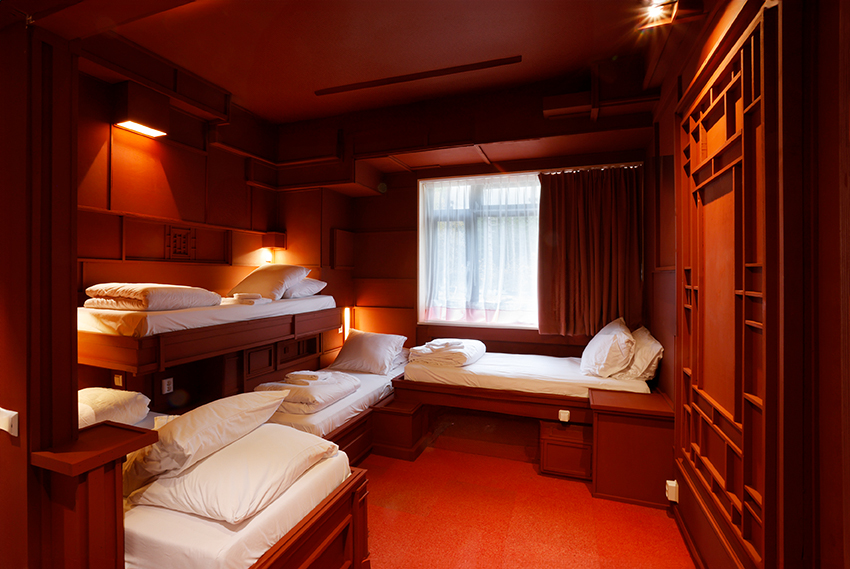 Hotelkamer van kunstenaar Jonathan Straatman met roodgeverfde vloer, plafond en muren met daarop driedimensionale geometrische vormen. In de kamer staan vier rode eenpersoonsbedden met wit beddengoed, die op verschillende hoogtes en verschillende kanten op staan.