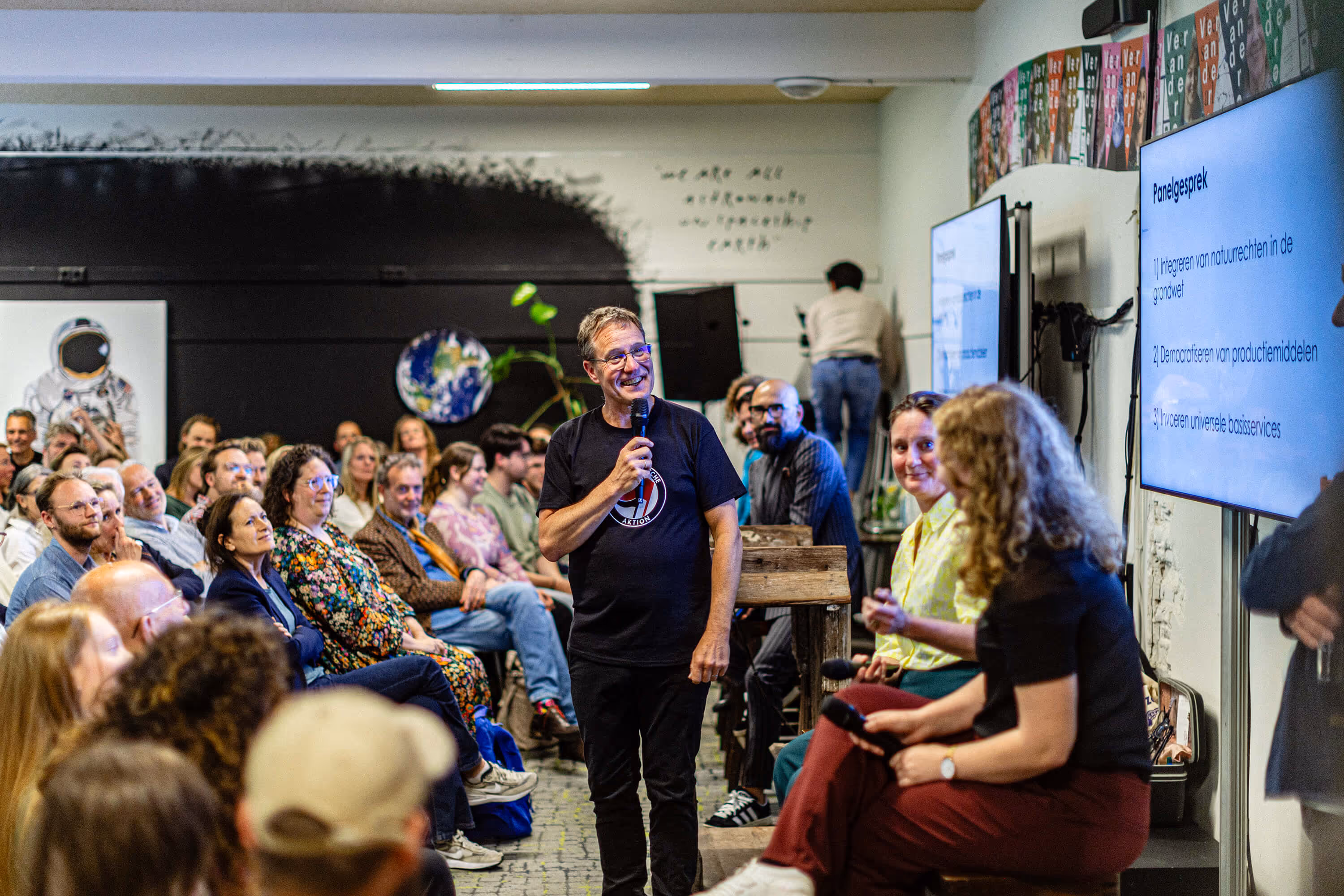 Mannelijke spreker staat glimlachend voor een publiek van tientallen mensen. Hij heeft een microfoon vast en kijkt naar een diascherm waarop een presentatie wordt getoond. Naast hem zitten nog twee vrouwelijke sprekers met microfoon in de hand.