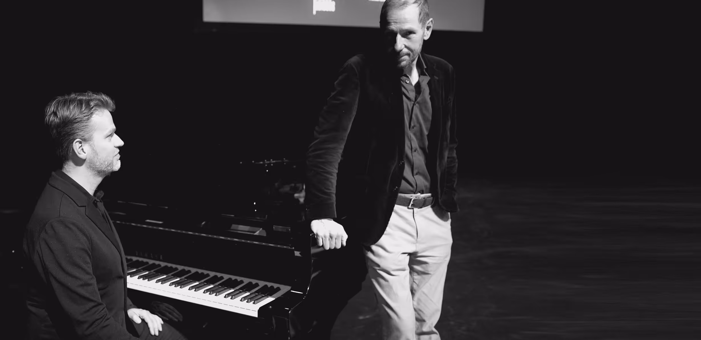 Roek Lips staat naast en Bas Bons zit achter een piano op het podium.