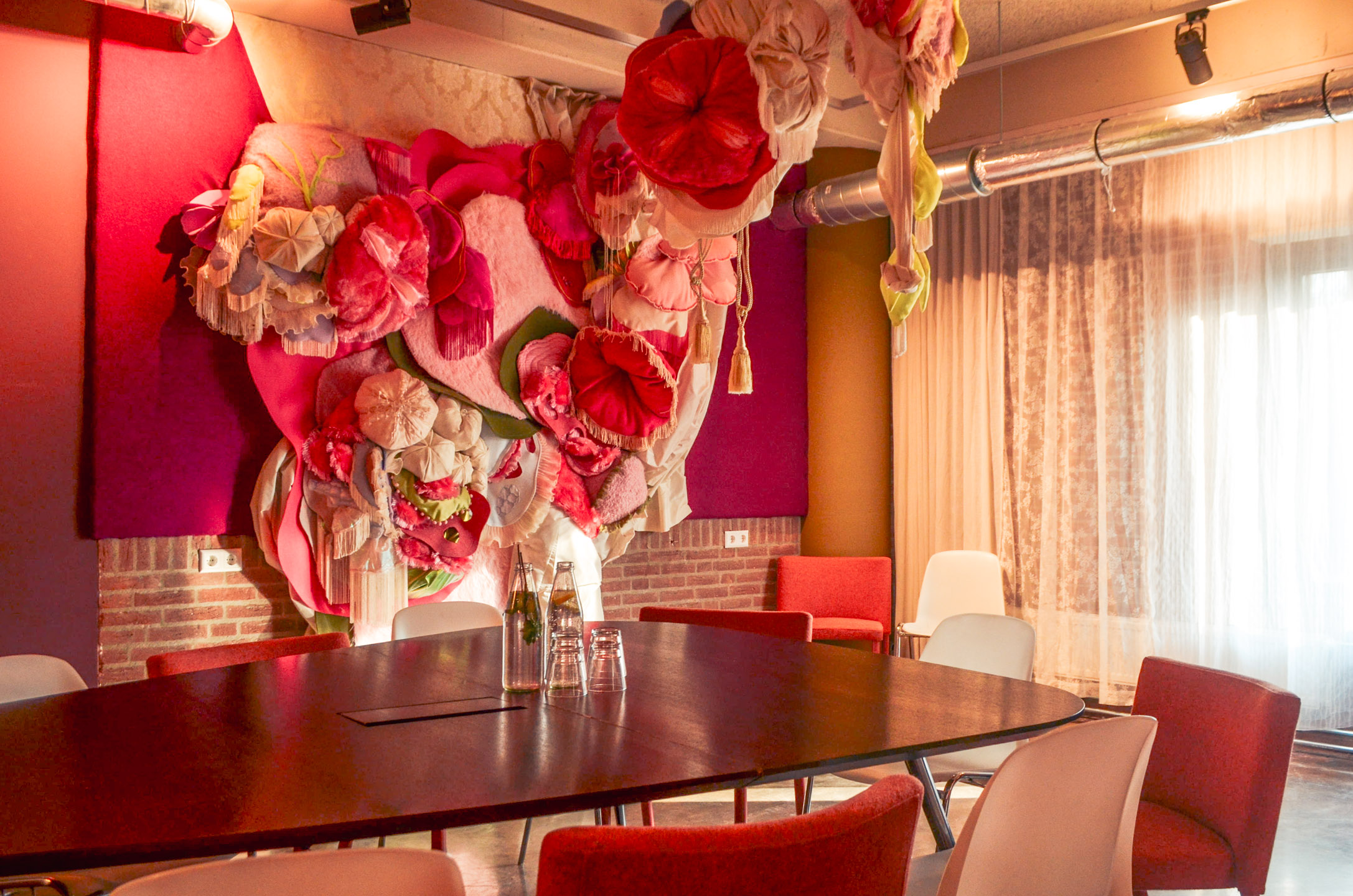 Detail een abstract driedimensionaal kunstwerk van zachte textiel met meerdere kleuren en structuren, at aan de muur hangt in De Fijne Kamer van het Ongemak bij De Groene Afslag.