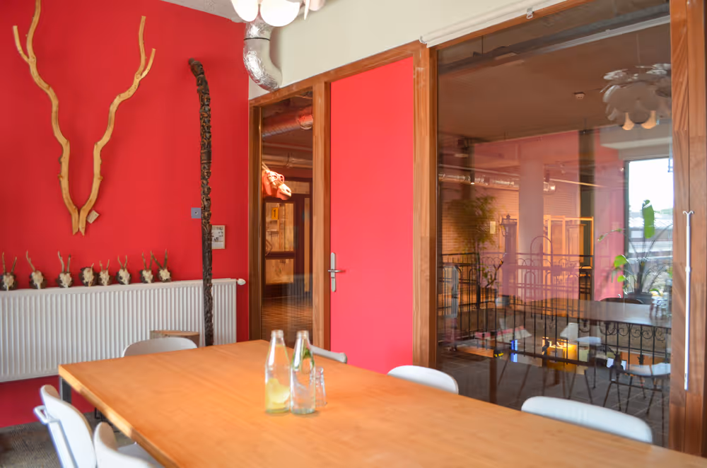 Vergaderruimte De Roze Bril bij De Groene Afslag. Grote houten tafel met lichtgrijze stoelen eromheen in een donkerroze geschilderde ruimte met grote ramen aan weerszijden. 