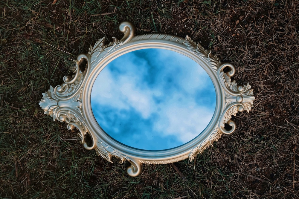 Spiegel die op gras ligt waardoor je de blauwe lucht ziet weerspiegeld.