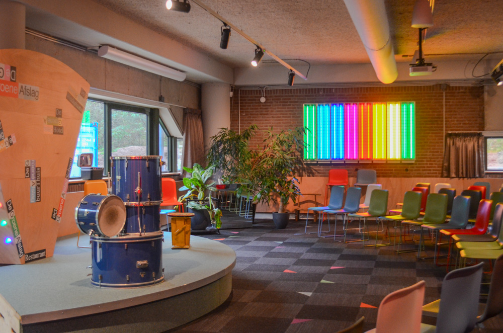  Overzichtsfoto van de Grootste Zaal bij De Groene Afslag. Rijen stoelen in theater opstelling en een groot diascherm boven een klein podium.