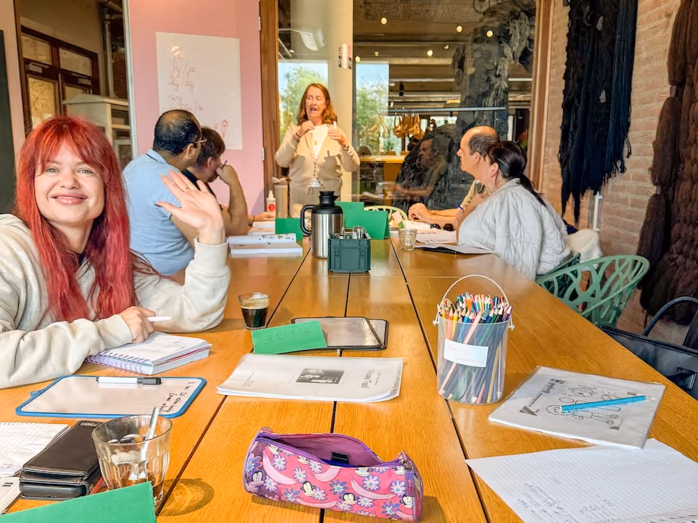 Een groep mensen rond een tafel, de voorsta vrouw zwaait vriendelijk naar de camera.