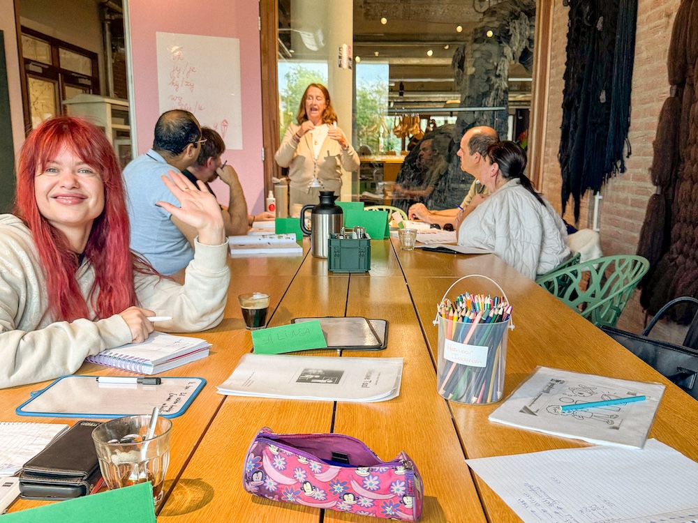 Een groep mensen rond een tafel, de voorsta vrouw zwaait vriendelijk naar de camera.