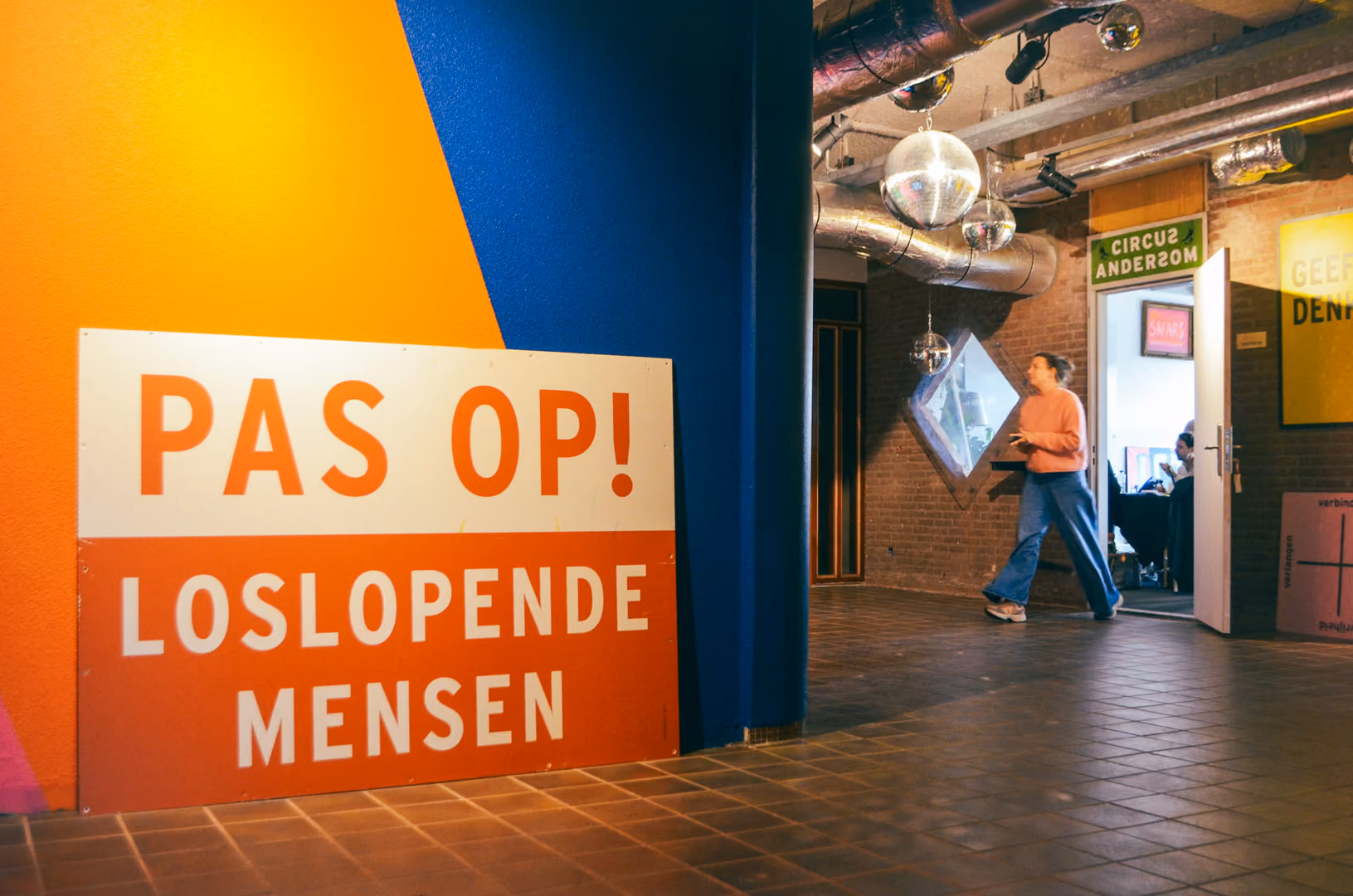 Een bord met de tekst 'PAS OP! LOSLOPENDE MENSEN' op een oranje en blauwe muur in een ruimte waar een vrouw in een roze trui en blauwe broek langsloopt.