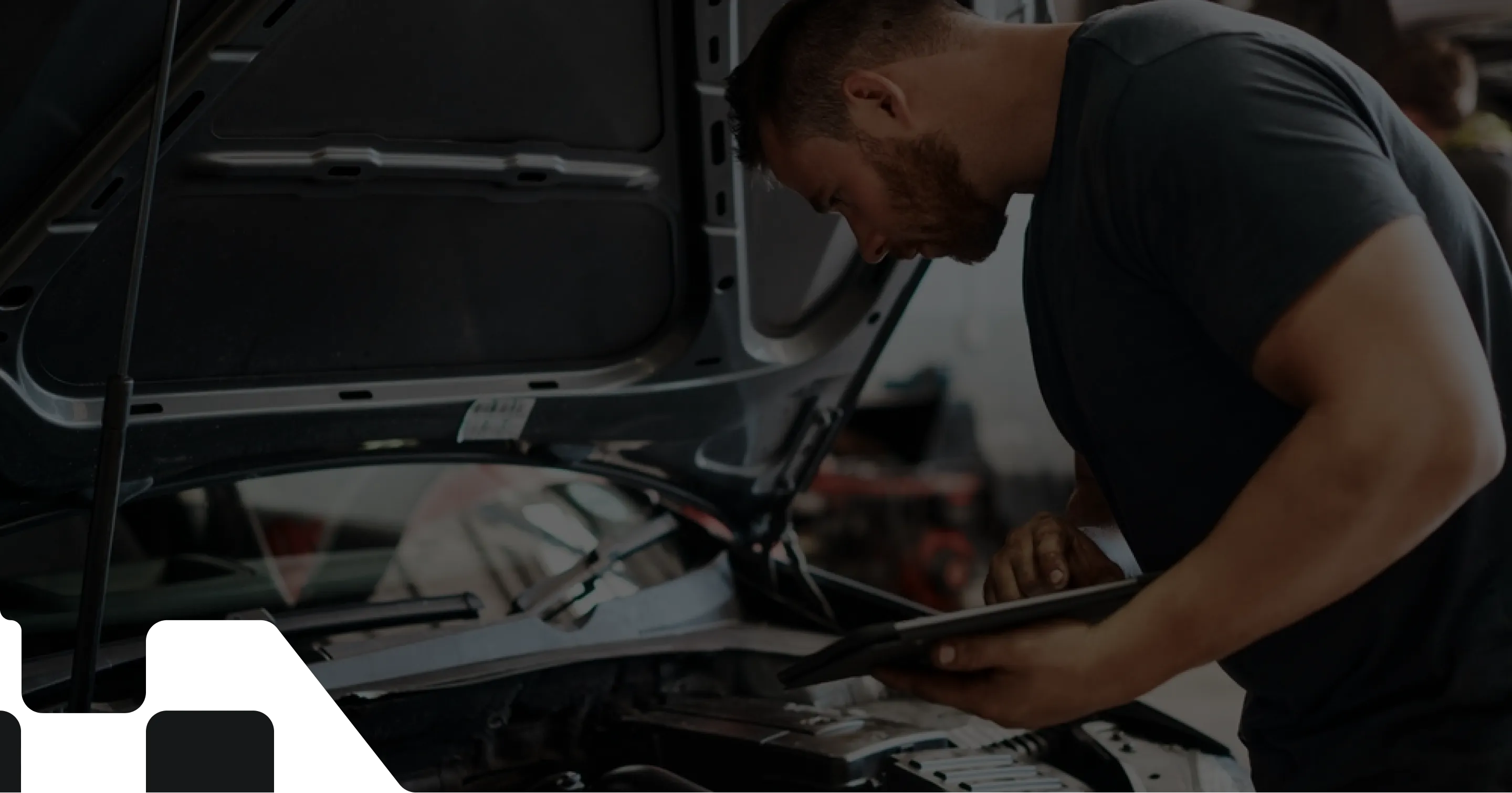 A male mechanic with a beard, wearing a dark t-shirt, leaning over an open car hood while using a tablet to inspect the engine in a workshop setting.