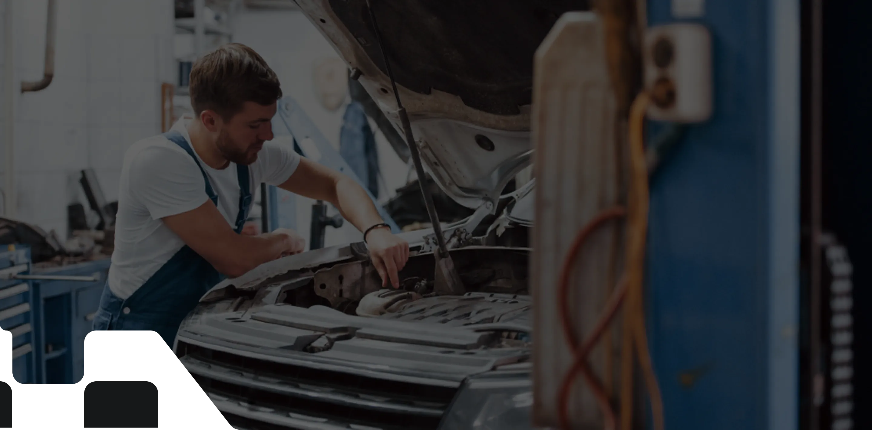 A male mechanic with short brown hair and a beard, wearing a white t-shirt and blue overalls, is leaning over the open hood of a car and working on the engine. The garage setting includes visible tools, automotive equipment, and another car lift in the background.