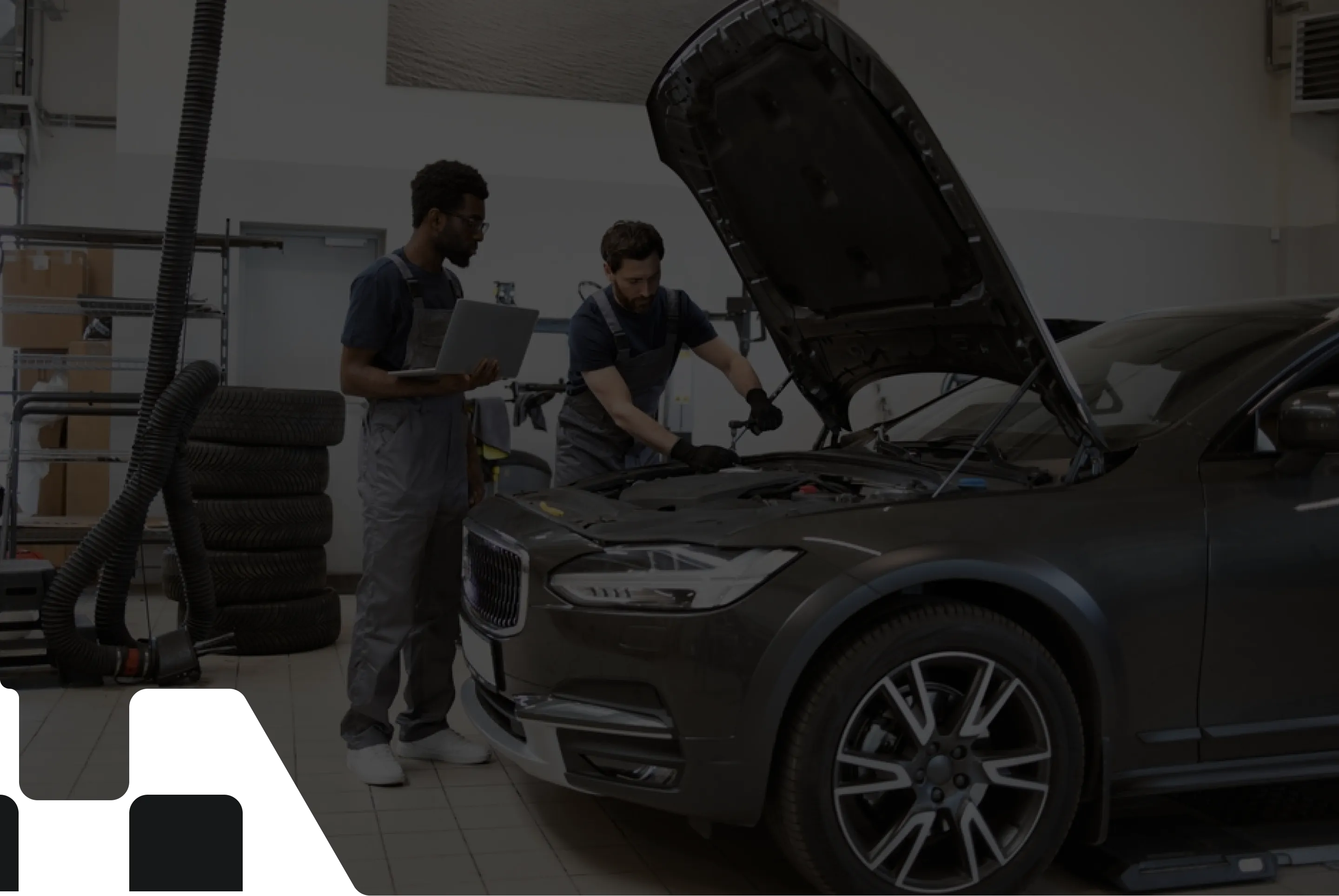 Two auto mechanics inspecting a car with its hood open in a garage; one mechanic uses a laptop while the other examines the engine.