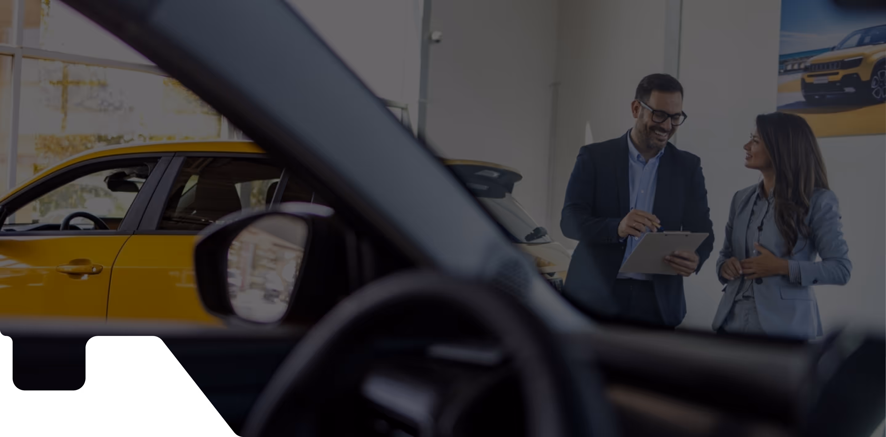 Interior view from inside a car at a dealership, with the steering wheel and side mirror in the foreground. In the background, a man in a suit holding a clipboard is conversing with a woman in business attire, both standing near a yellow car. A poster of a yellow vehicle is visible on the wall behind them. Bright windows let in natural light, highlighting the sales environment.