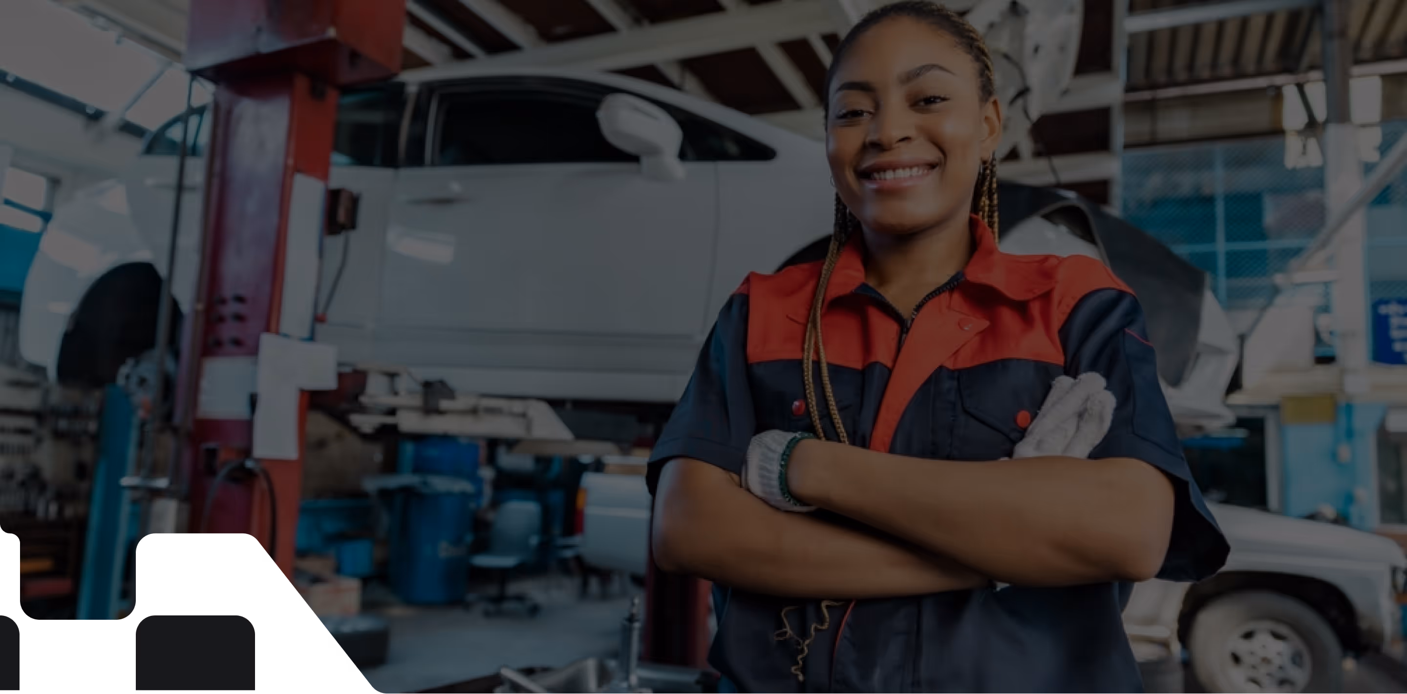 Confident woman with dark skin and braided hair standing in a car repair shop, smiling with arms crossed. She is wearing a work uniform with red and navy blue colors, and gloves. Behind her, a car is lifted on a hydraulic lift and various tools and equipment are visible, highlighting the workshop environment.