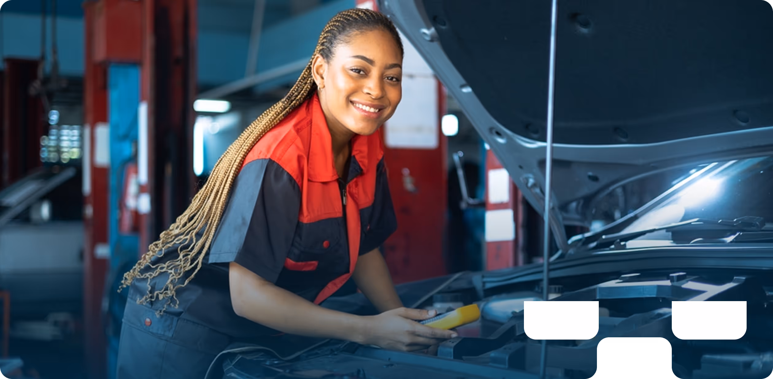 Woman mechanic with braided hair smiling while leaning over an open car hood in an auto repair shop. She is wearing a red and black uniform and holding a tool. The background shows car repair equipment and the interior of the garage.