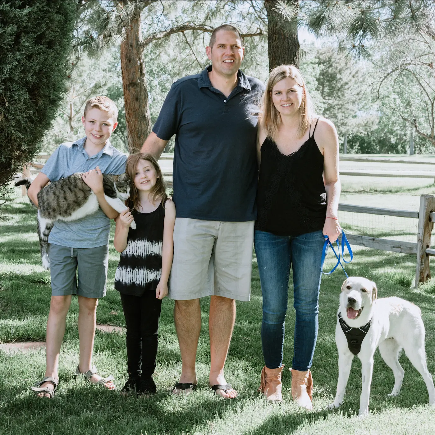 Julie, a Colorado Wealth Group client, stands with her smiling family, including two kids, a cat, and a dog, in a sunny park setting, symbolizing financial stability and a fulfilling lifestyle.