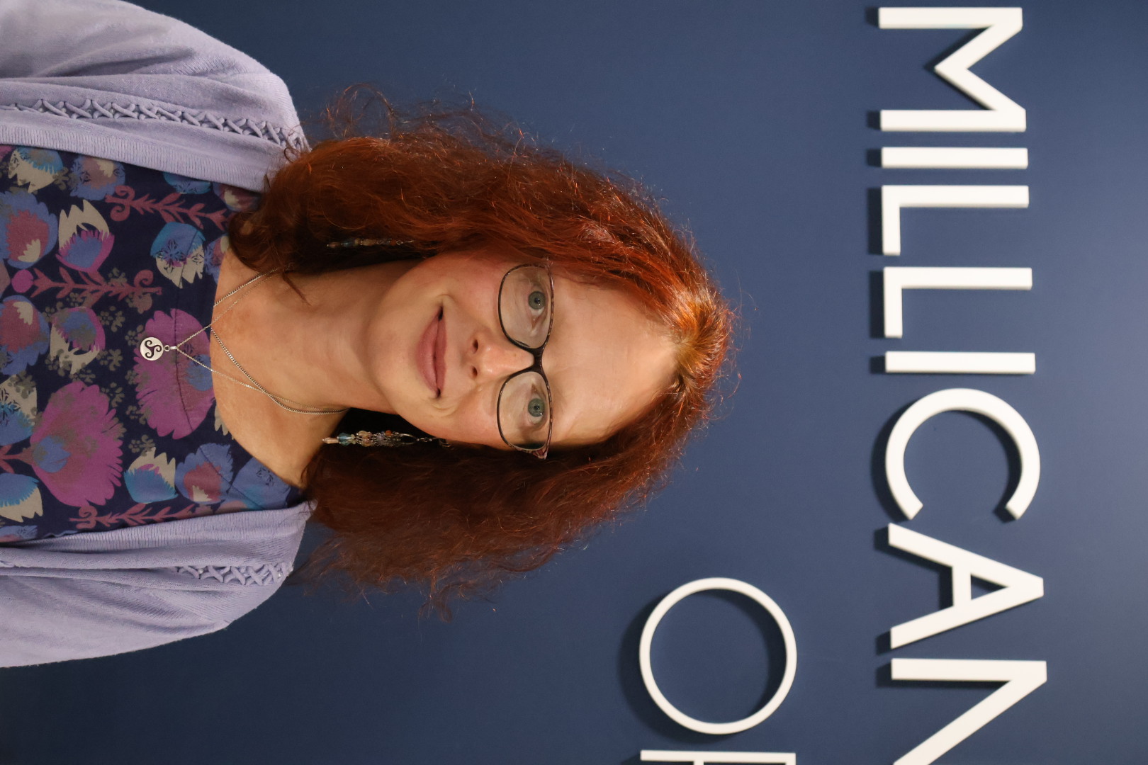 Woman with curly red hair and glasses posing in front of Millican sign
