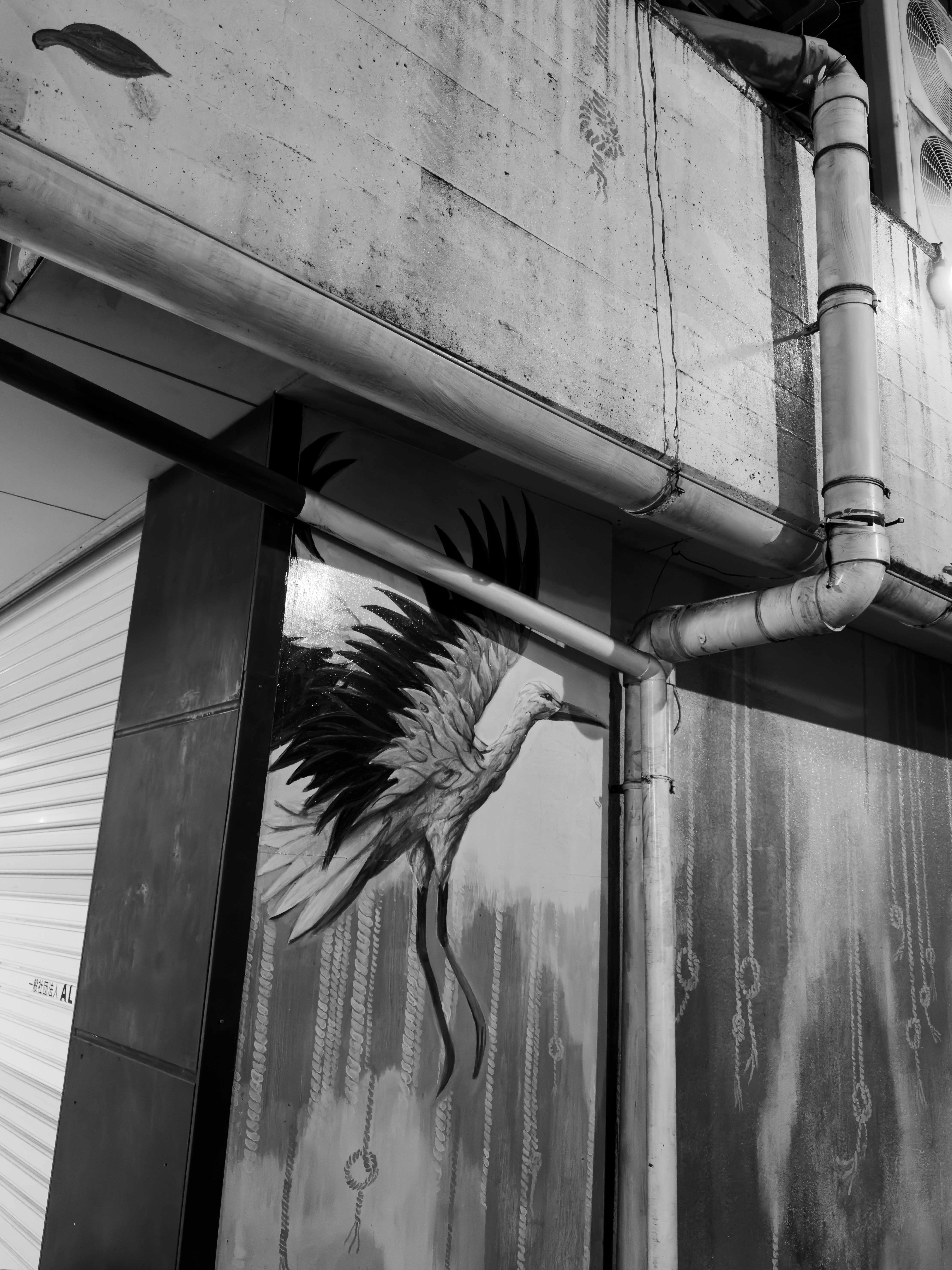 Black and white photo of a mural depicting a bird with outstretched wings on a building wall with pipes and chains.