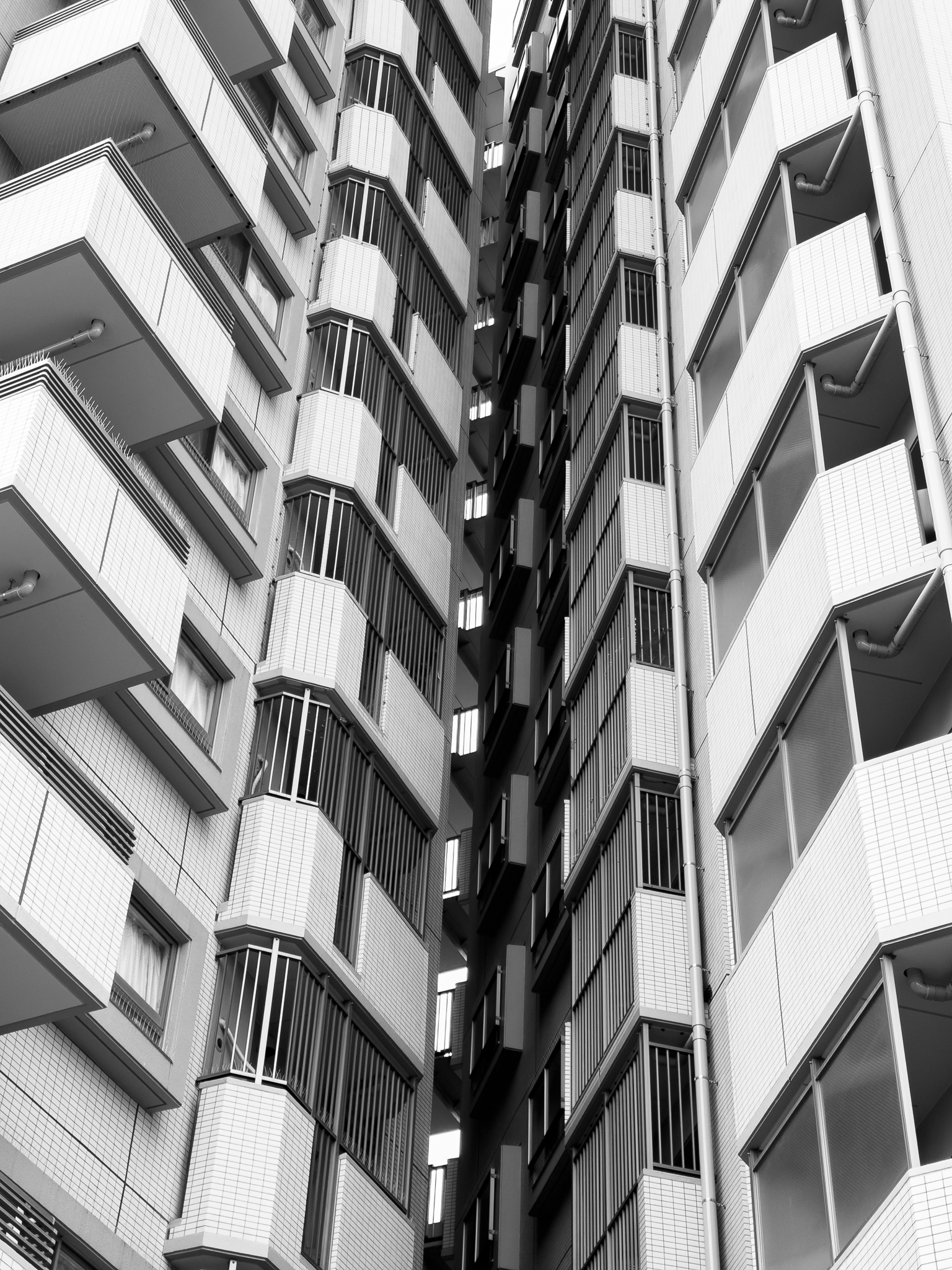 Black and white photo of two tall apartment buildings with balconies and narrow space between them.