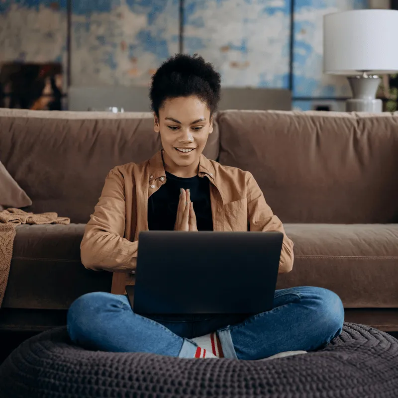 Pretty coloured woman sitting in front of the sofa on her laptop looking positive