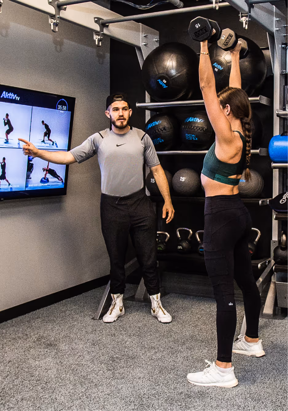 Fitness trainer instructing a woman lifting dumbbells overhead in a gym with exercise balls and kettlebells in the background.