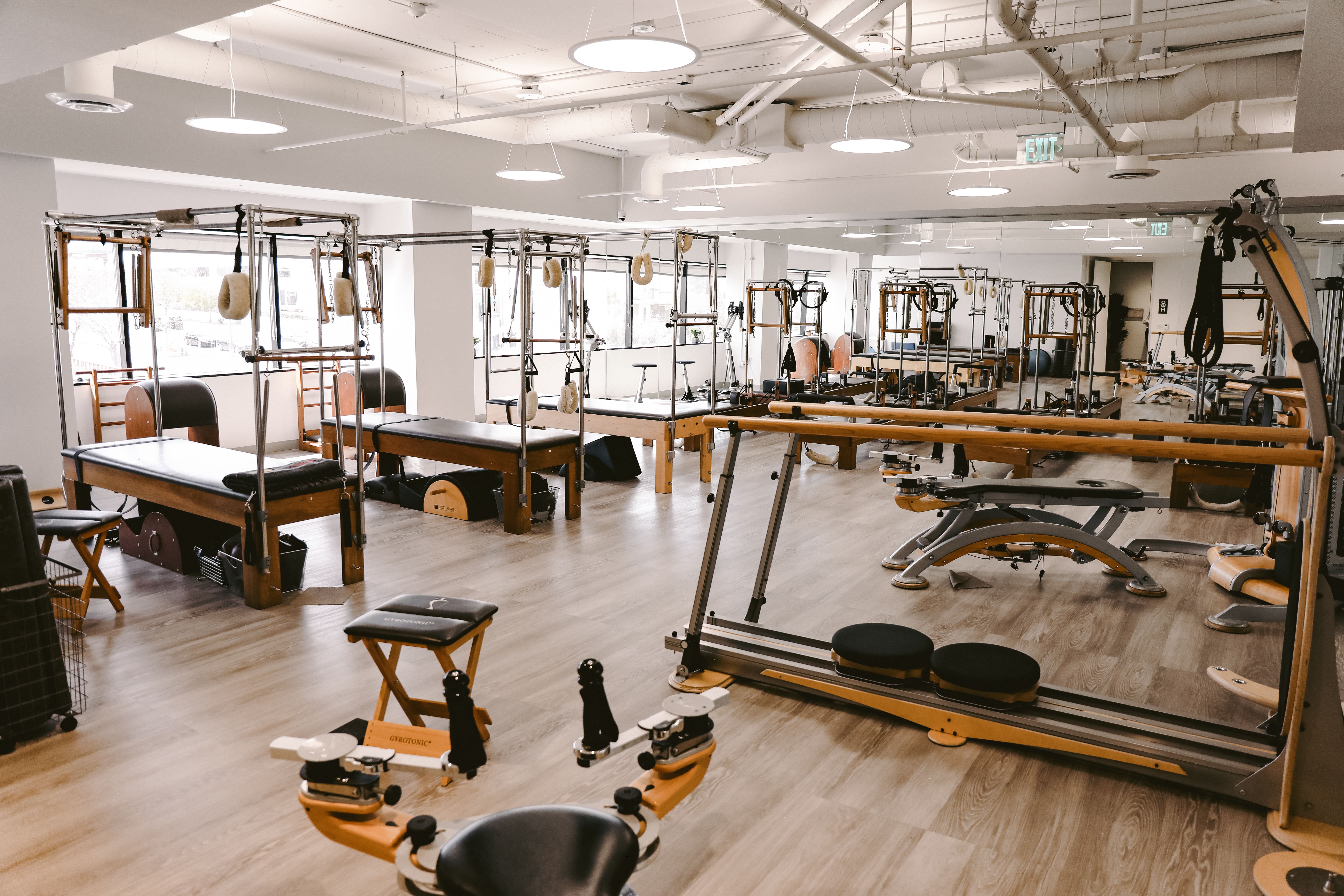 Bright Pilates studio with wooden reformer machines, chairs, and fitness equipment on light wood flooring.
