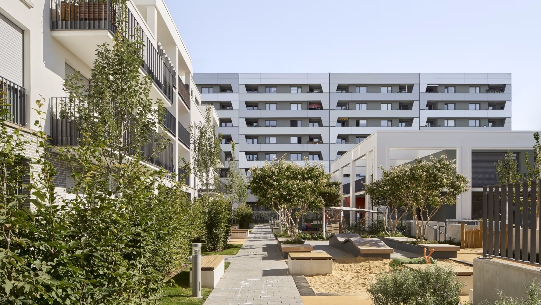 Moderner Wohnhof mit beblühter Sandspielfläche, Bänken und gepflegten Büschen zwischen mehrstöckigen Wohngebäuden.