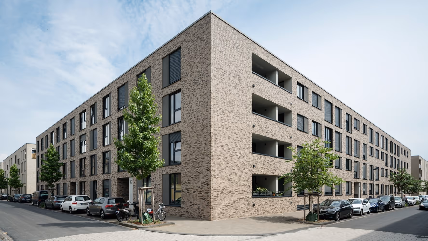 Moderner, dreistöckiger Backsteinwohnblock mit Bäumen und geparkten Autos an beiden Straßenseiten.