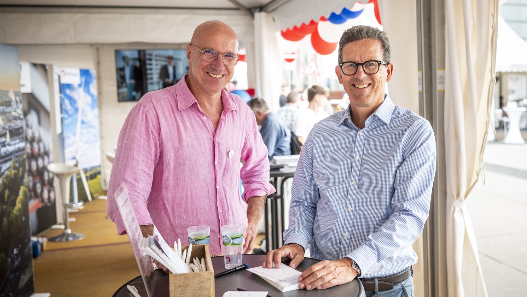 Zwei lächelnde Männer an einem Stehtisch mit Informationsmaterial unter einem Zelt mit rot-weiß-blauer Dekoration.