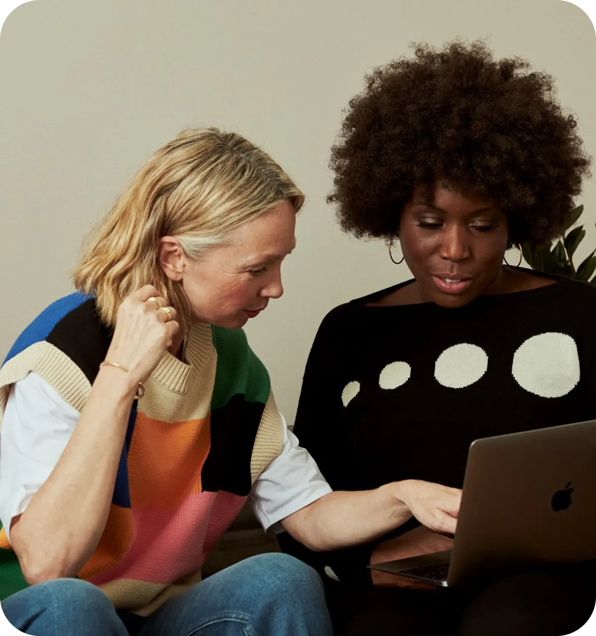 Two women sitting together and looking at a laptop screen, one pointing at the display while the other focuses on it attentively.
