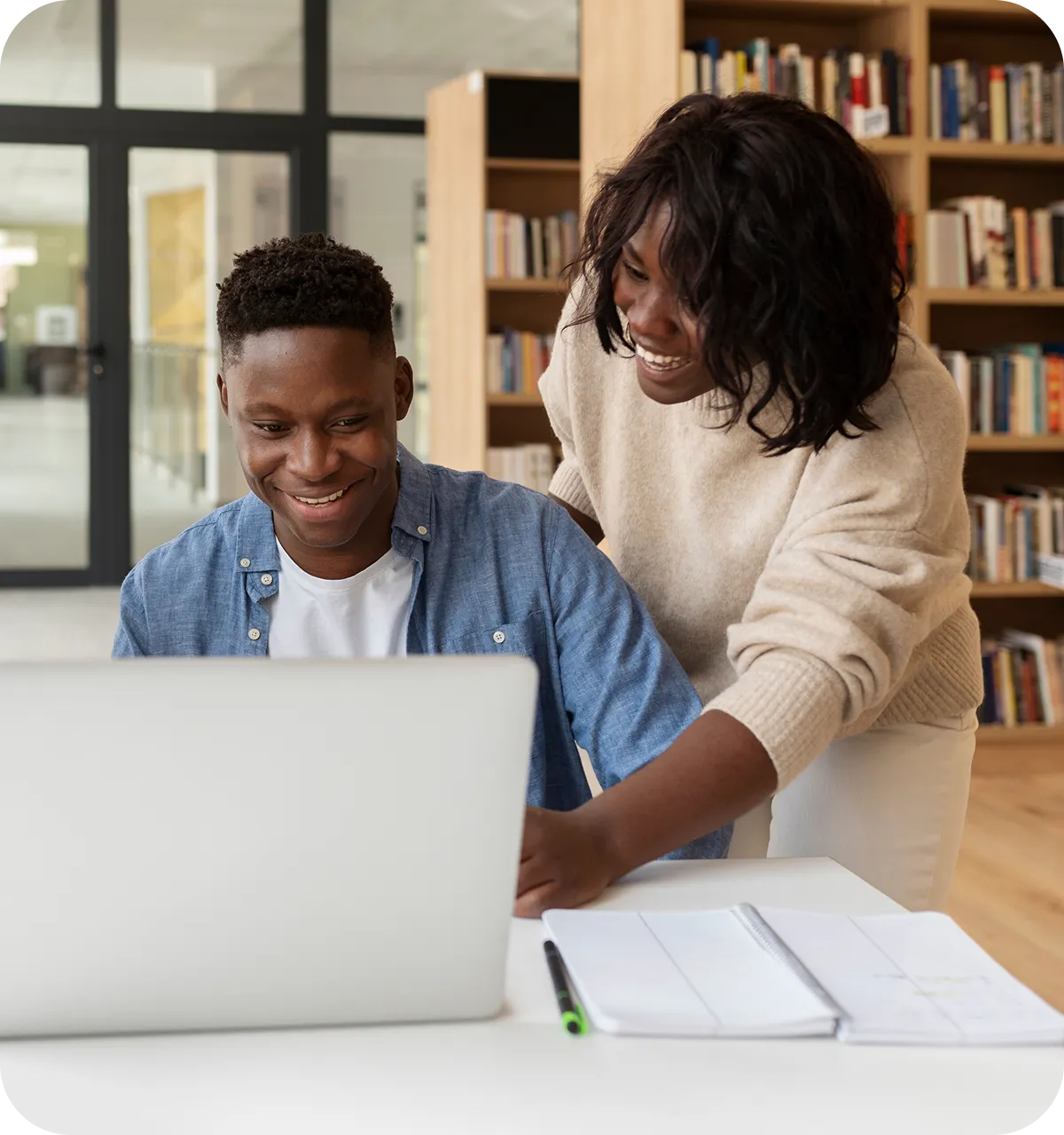 Two students smiling and working together at a laptop in a modern library, with notebooks and bookshelves in the background.
