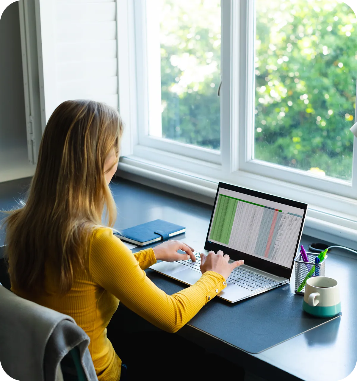 Person working on a spreadsheet at a laptop near a bright window, with a notebook, pen holder, and mug on the desk.
