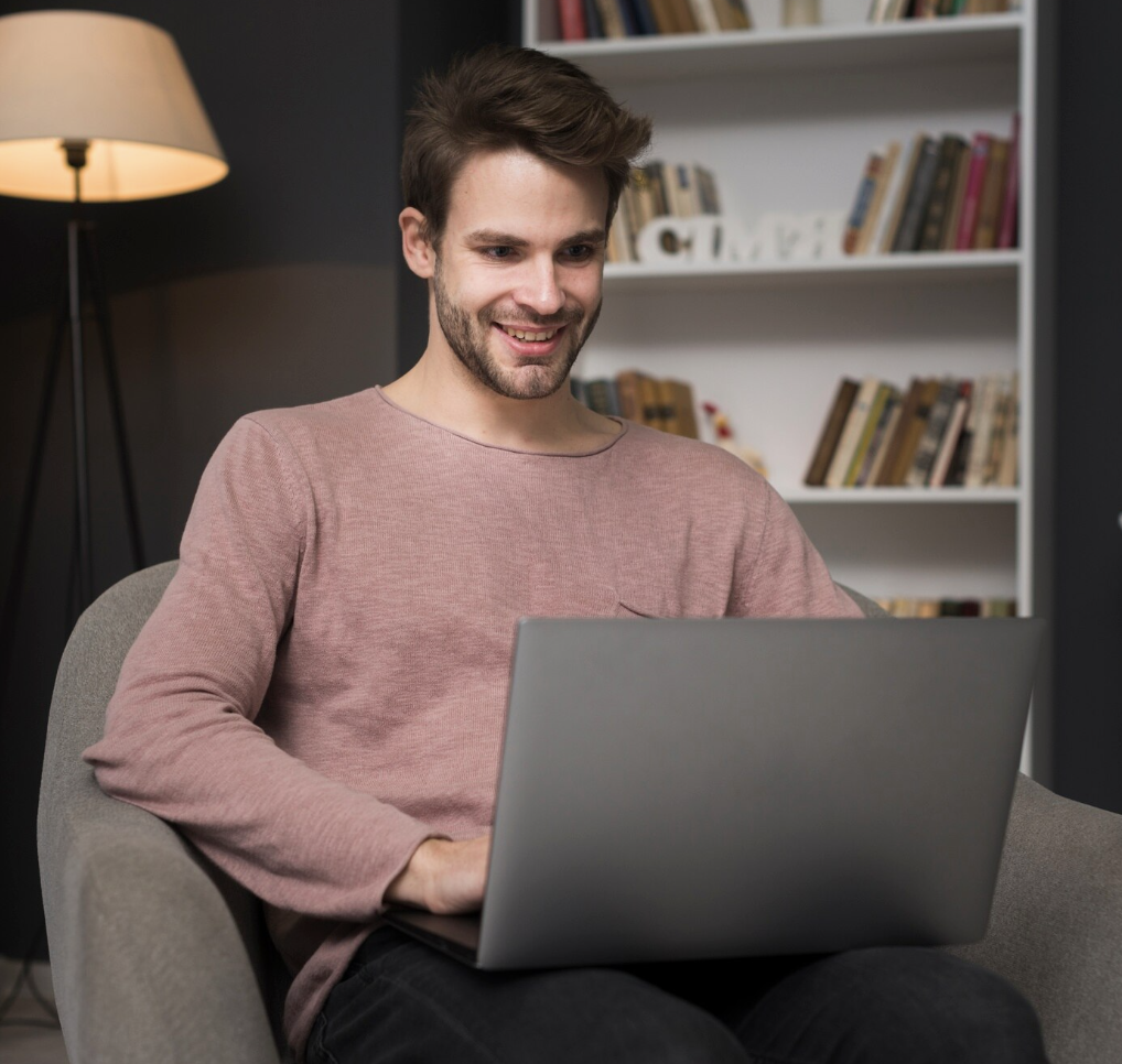 Young person with curly hair working on a laptop in a library or study area, with bookshelves and a notebook visible in the background.
