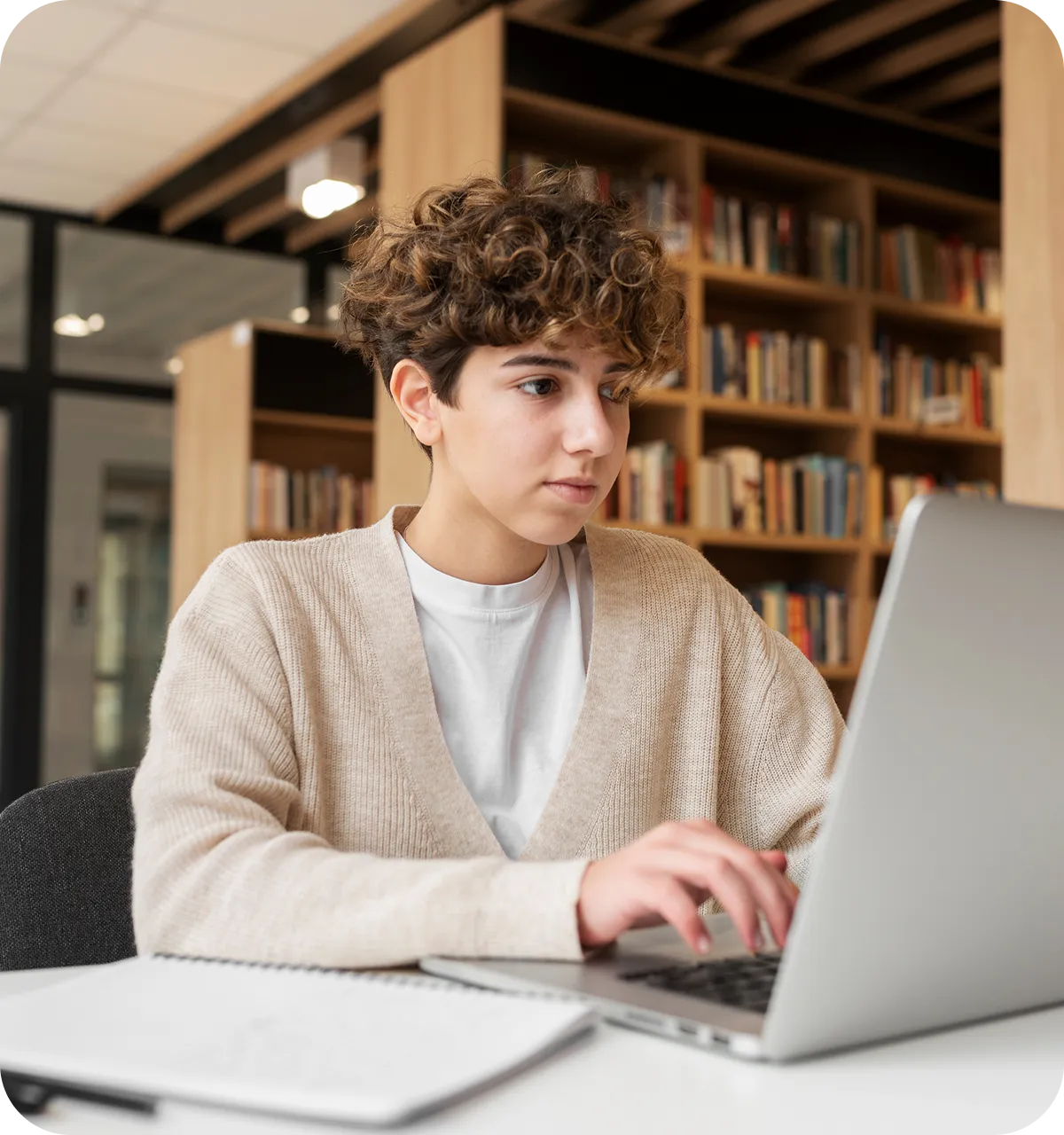 Young person with curly hair working on a laptop in a library or study area, with bookshelves and a notebook visible in the background.
