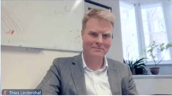 Thies Lindenthal on a video call, smiling in a gray blazer with a whiteboard and window in the background.