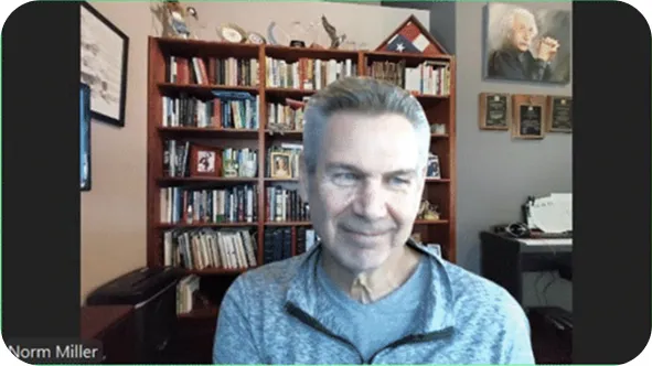 Norm Miller during a video call, smiling in front of a large bookshelf and framed photos in a home office.