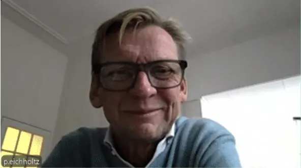 Piet Eichholtz smiling during a video call, wearing glasses and a blue sweater in a softly lit room.