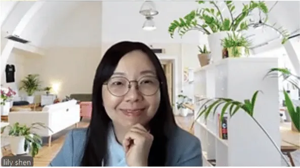 Lily Shen smiling during a video call, seated in a bright room with plants and modern furniture in the background.