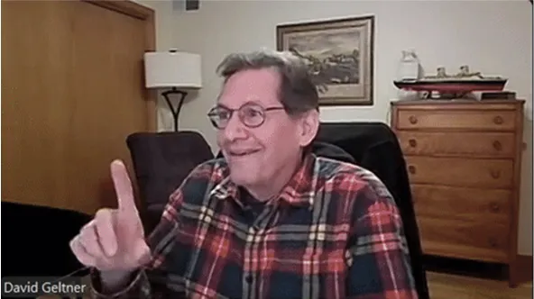 David Geltner smiling during a video call, wearing a plaid shirt and raising his index finger.
