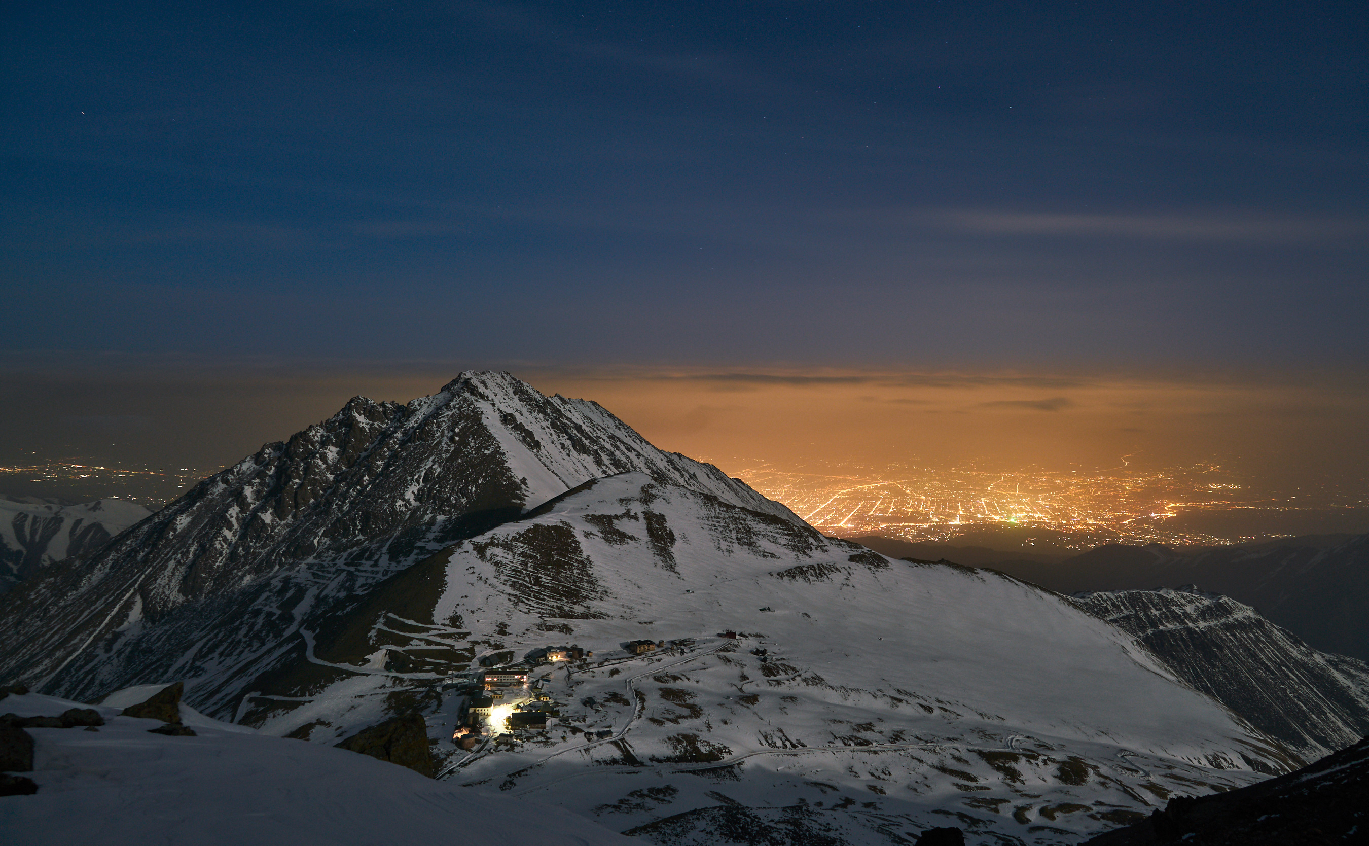 Night view of Almaty from a snow-covered mountain, with city lights glowing in the distance under a deep blue sky.
