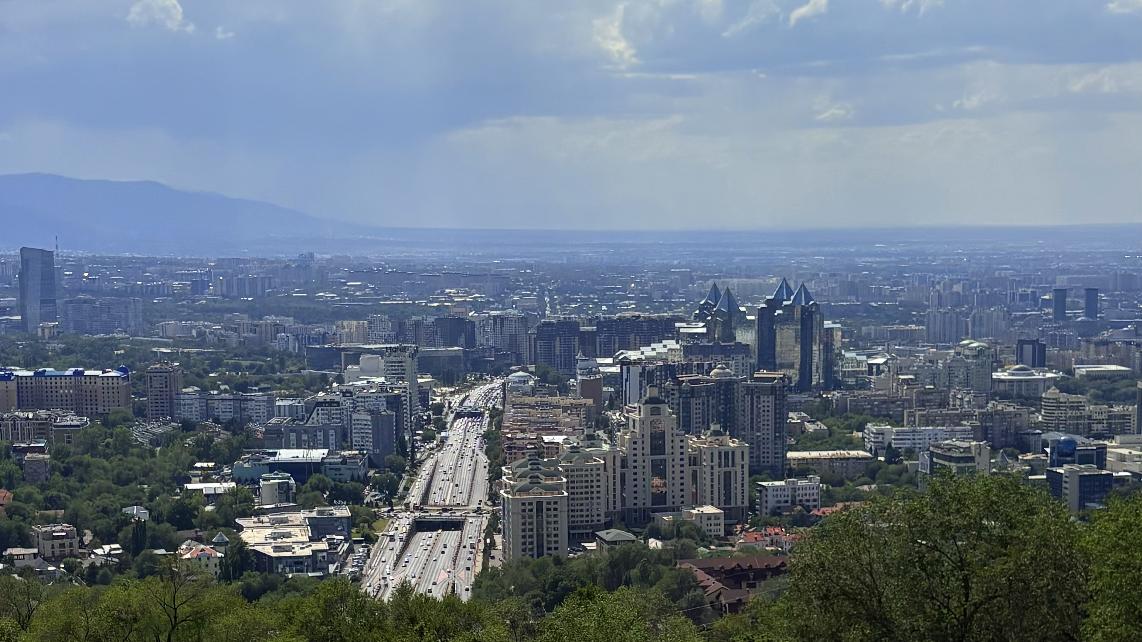 Panoramic skyline of Almaty at sunset, featuring modern buildings in the foreground and the Tien Shan mountains in the background under a clear sky.