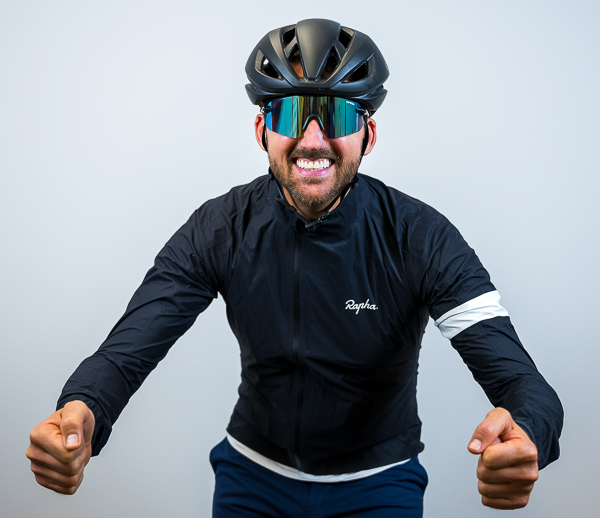 Steven Harp, Founder and CEO of Colorado Wealth Group, dressed in full cycling gear. He is smiling broadly while leaning forward in a racing pose, wearing a black Rapha cycling jacket, dark blue cycling shorts, a black aerodynamic helmet, and reflective wraparound sunglasses. The light gray background emphasizes his energetic and athletic stance.