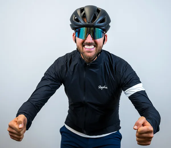 Steven Harp, Founder and CEO of Colorado Wealth Group, dressed in full cycling gear. He is smiling broadly while leaning forward in a racing pose, wearing a black Rapha cycling jacket, dark blue cycling shorts, a black aerodynamic helmet, and reflective wraparound sunglasses. The light gray background emphasizes his energetic and athletic stance.